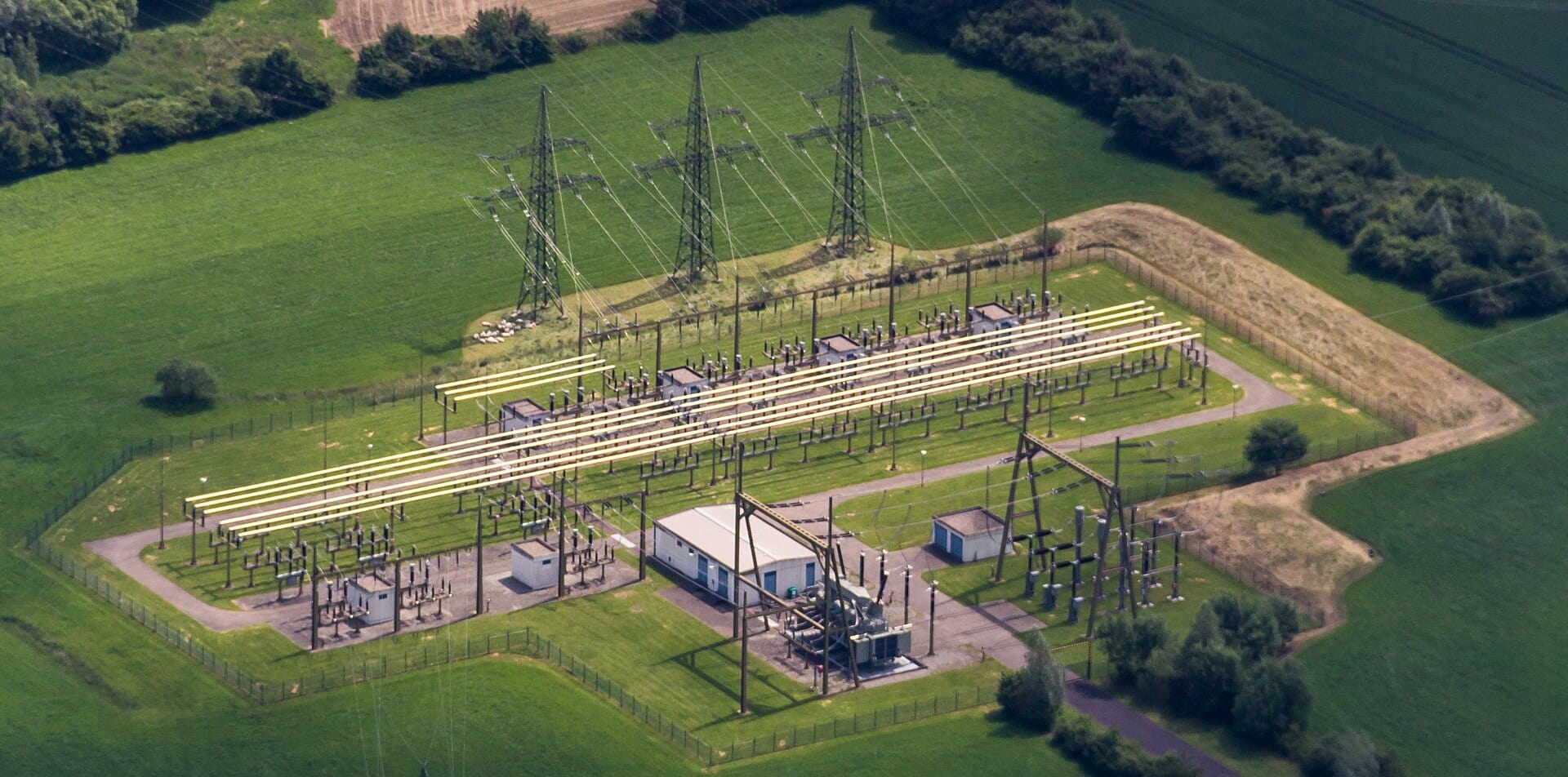 An aerial view of a transmission substation, showing parallel busbars running the length of the yard between rows of switchgear, with overhead lines arriving from three pylons.